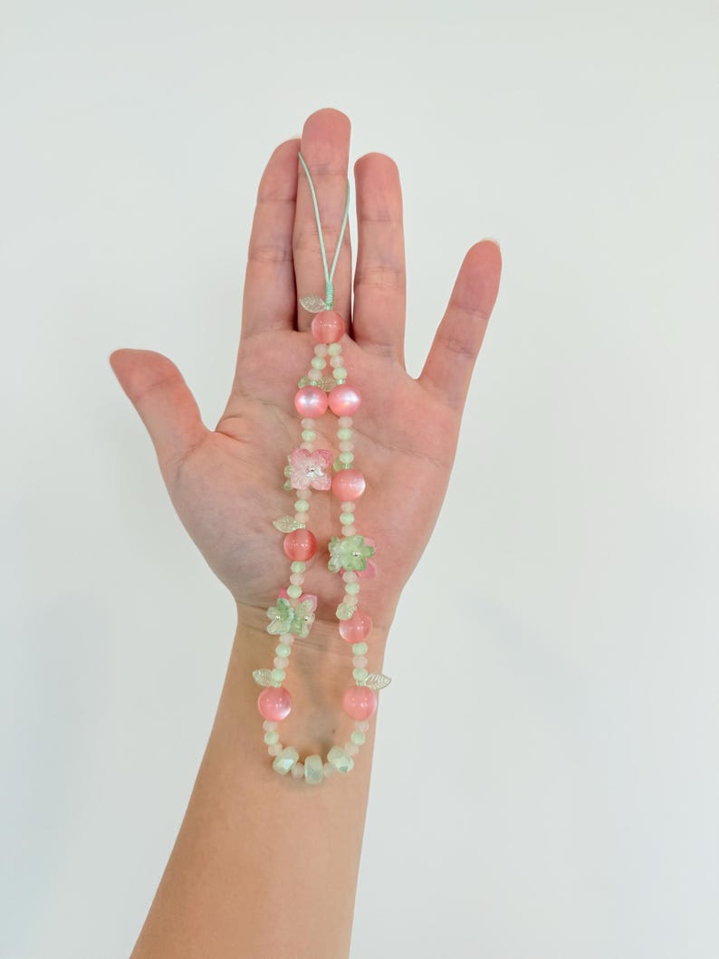 May include: A beaded necklace featuring pink and green beads, with flower and leaf accents. The necklace is held against a white background. The beads are a mix of round and flower-shaped, with a light green cord.