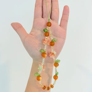May include: A beaded phone charm featuring orange-colored beads resembling oranges, interspersed with green leaf beads and small pink flower beads. The charm is strung on a green cord.