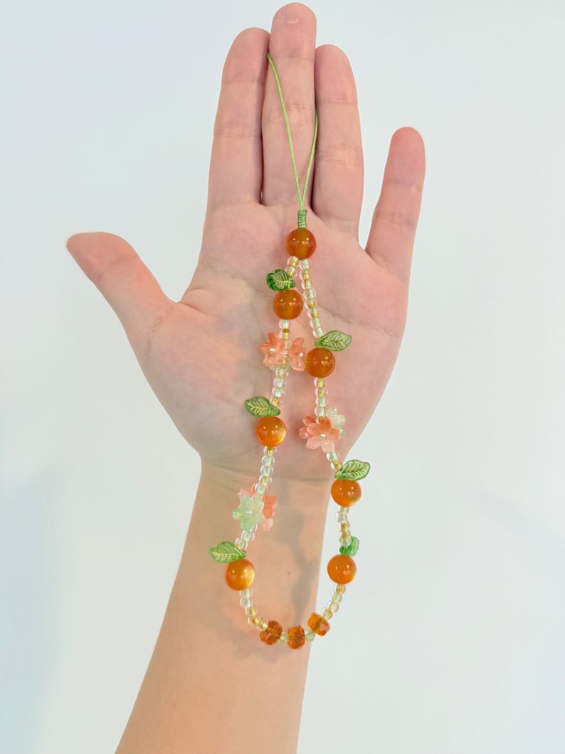 May include: A beaded phone charm featuring orange-colored beads resembling oranges, interspersed with green leaf beads and small pink flower beads. The charm is strung on a green cord.