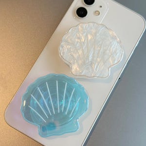Peut inclure: Deux supports de téléphone en forme de coquillage sont fixés à un smartphone blanc. L'un est blanc avec une finition nacrée et une bordure dorée, l'autre est bleu translucide avec des lignes blanches.