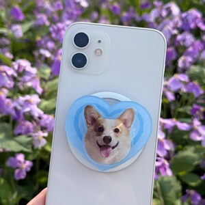 May include: A white smartphone with a light blue heart-shaped phone grip featuring a smiling corgi dog. The phone grip is attached to the back of the phone. The background is a blurred view of purple flowers.