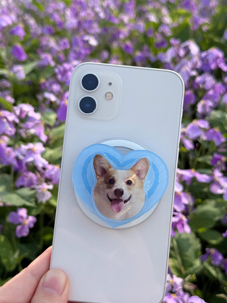 May include: A white smartphone with a light blue heart-shaped phone grip featuring a smiling corgi dog. The phone grip is attached to the back of the phone. The background is a blurred view of purple flowers.