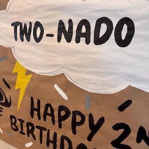 Puede incluir: Una pancarta de papel marrón con pintura negra y blanca. La pancarta dice "TWO-NADO HAPPY BIRTHDAY 2".