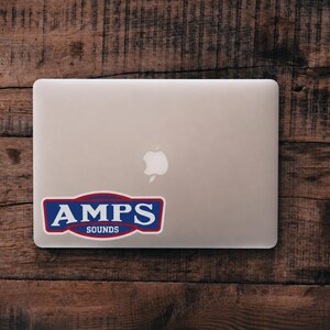 以下が含まれることがあります： 青い背景、赤い縁取り、そしてセリフ体で白い文字のAMPS SOUNDSと書かれた長方形のステッカーが貼られたシルバーのノートパソコン。ステッカーは、中央にあるAppleロゴの下、ノートパソコンの蓋の左下に配置されています。