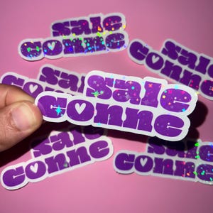 Peut inclure: Une collection d'autocollants violets portant le texte "sale conne" dans une police grasse et arrondie. Chaque autocollant comprend une petite icône de cœur blanc entre les mots et présente une finition holographique aux reflets étincelants.