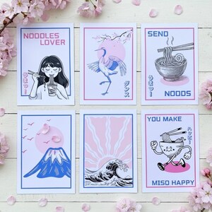 Peut inclure: Six illustrations sur papier avec des motifs roses et bleus. Les dessins comprennent une femme mangeant des nouilles, une grue, un bol de nouilles, une montagne, une vague et un bol de nouilles avec un visage. Le texte comprend "NOODLES LOVER", "SEND NOODS" et "MISO HAPPY".