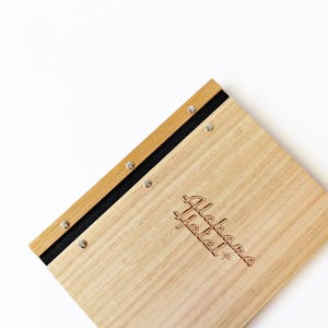 Timber compendium rests on a white background. The folder is made of Tasmanian oak, with black binding and 6 nickel screws. The cover is engraved with the logo of boutique hotel.