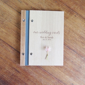 A folder with wood Tasmanian oak covers sits on a darker timber table. The cover is engraved with Our Wedding Cards and personal details. A pink gum blossom sits on the book cover.