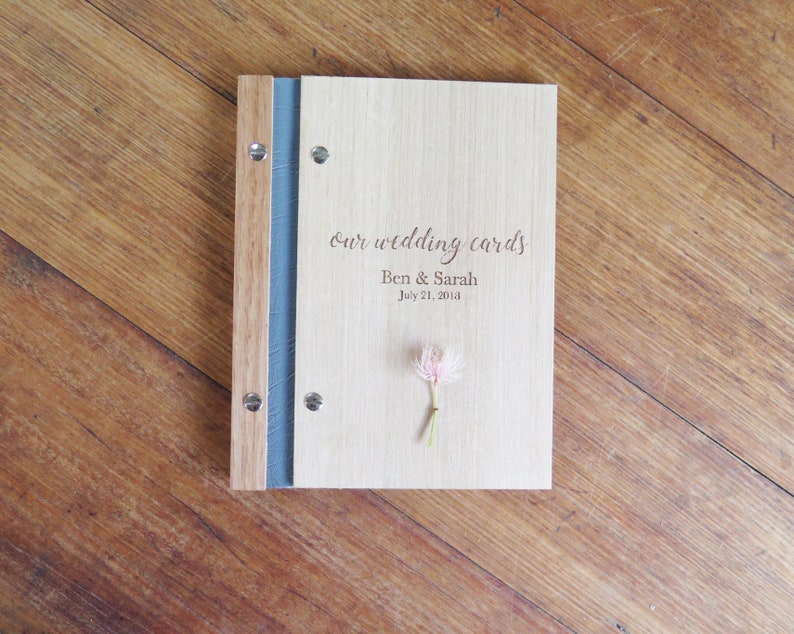 A folder with wood Tasmanian oak covers sits on a darker timber table. The cover is engraved with Our Wedding Cards and personal details. A pink gum blossom sits on the book cover.