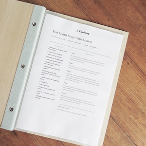 A recipe folder made with timber covers lies open on a wooden table. The plastic sleeve inserts hold printed recipe pages.
