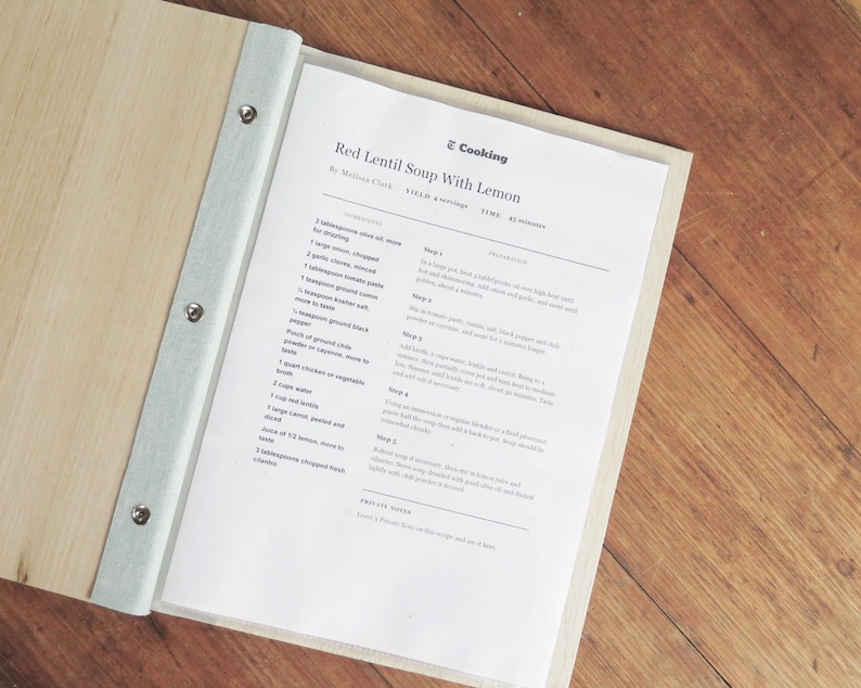 A recipe folder made with timber covers lies open on a wooden table. The plastic sleeve inserts hold printed recipe pages.