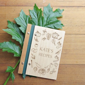 A recipe book with Tasmanian oak covers sits on a darker timber table. Green foliage is arranged around the book. The cover is engraved with a personal name and line drawings of various vegetables.