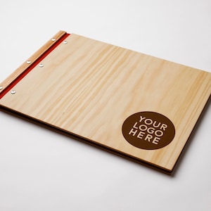 A large timber portfolio with pine  covers lies on a white background. The folder indicates a spot available for custom logo engraving. The folder has red vegan leather binding and 6 nickel screws.