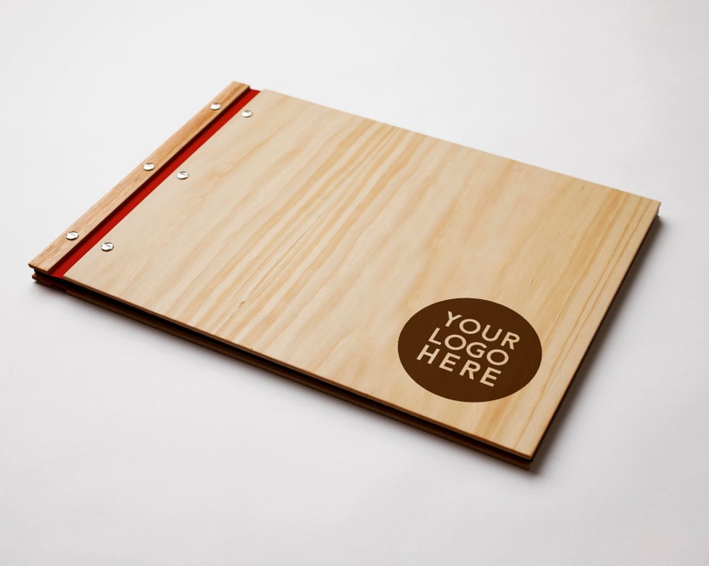 A large timber portfolio with pine  covers lies on a white background. The folder indicates a spot available for custom logo engraving. The folder has red vegan leather binding and 6 nickel screws.