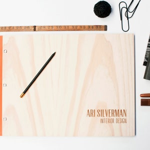 A professional portfolio with pine wood covers lies on a white desk surrounded by stationary.  The folder is engraved with a name. The folder has orange vegan leather binding and 6 nickel screws. It holds plastic sleeve inserts.