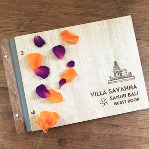 A hotel guestbook with Tasmanian oak wood covers lies on a timber bench top. The guest book is engraved with a logo for a villa in Bali. The book has grey vegan leather binding and purple and orange flower petals are strewn across the cover.