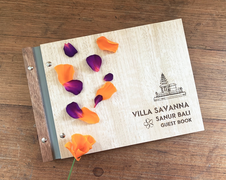 A hotel guestbook with Tasmanian oak wood covers lies on a timber bench top. The guest book is engraved with a logo for a villa in Bali. The book has grey vegan leather binding and purple and orange flower petals are strewn across the cover.