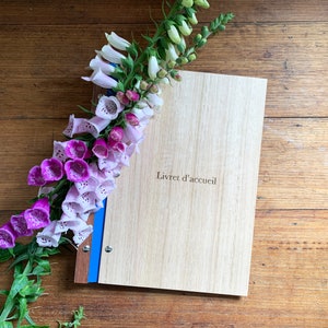 Timber compendium rests on a rustic wooden bench. Light and Dark pink foxglove flowers rest on the folder. The folder is made of Tasmanian oak, with blue binding and 6 nickel screws. The cover is engraved with Livret d'accueil