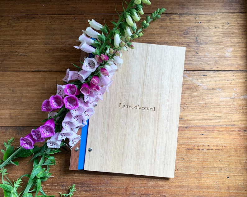 Timber compendium rests on a rustic wooden bench. Light and Dark pink foxglove flowers rest on the folder. The folder is made of Tasmanian oak, with blue binding and 6 nickel screws. The cover is engraved with Livret d'accueil