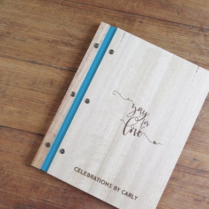 A celebrant book with wood Tasmanian oak covers sits on a darker timber table. The cover engraving is personalised, and the binding is a vibrant teal colour.