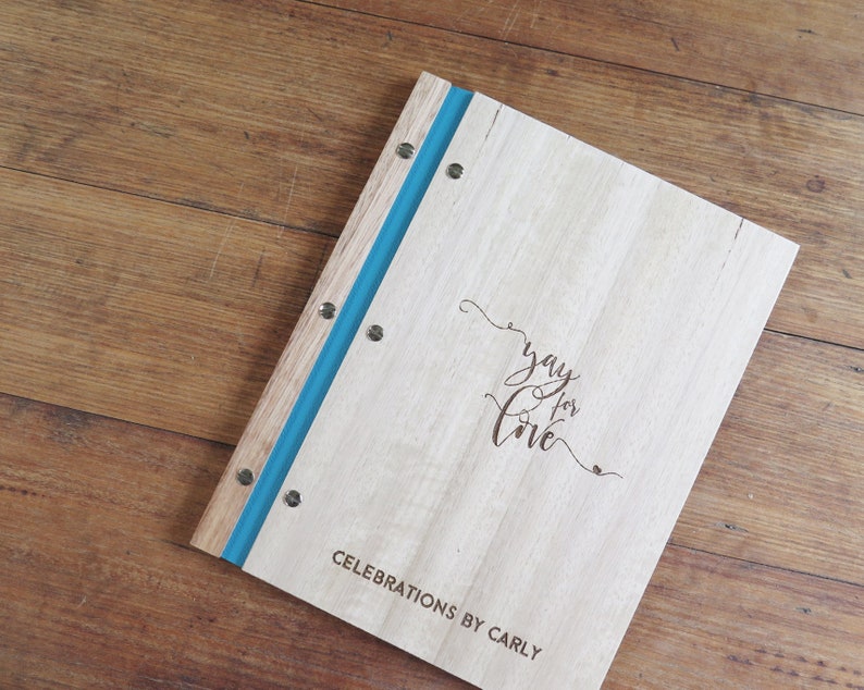 A celebrant book with wood Tasmanian oak covers sits on a darker timber table. The cover engraving is personalised, and the binding is a vibrant teal colour.