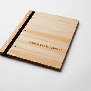 A professional portfolio with pine wood covers lies on a white background. The folder is engraved with a name. The folder has black vegan leather binding and 6 nickel screws. It holds plastic sleeve inserts.