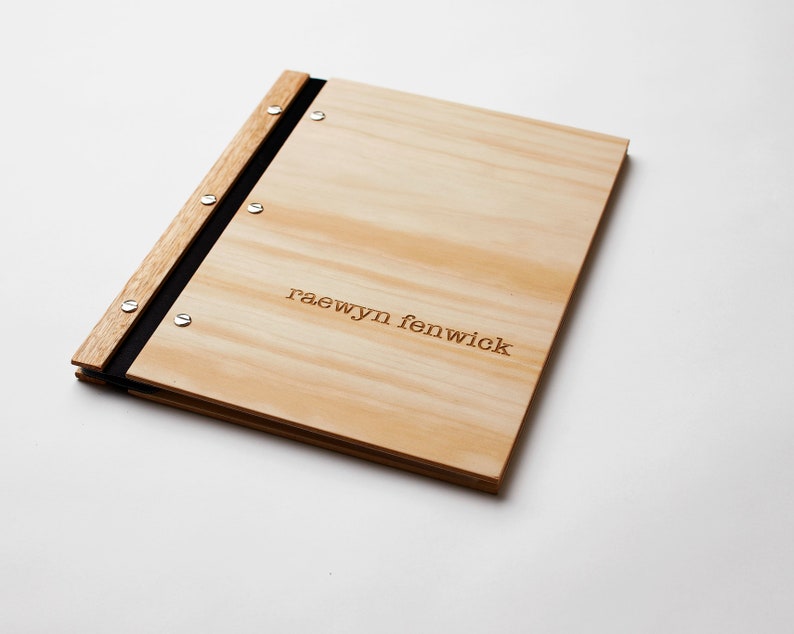 A professional portfolio with pine wood covers lies on a white background. The folder is engraved with a name. The folder has black vegan leather binding and 6 nickel screws. It holds plastic sleeve inserts.