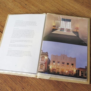 A timber A5 size compendium lies open on a wood table. The plastic sleeves display images of a boutique hotel, and a list of house rules.