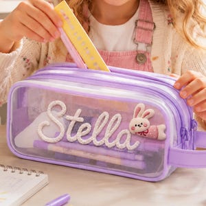 May include: A rectangular, transparent lavender pencil case with a zipper closure and a side handle. The front features the name Stella in white cursive script, accompanied by a small, plush, pink and white bunny character patch.