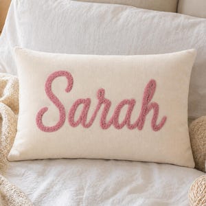 May include: Cream-colored rectangular pillow with the name "Sarah" embroidered in a script font with a textured, pink yarn. The pillow is placed on a white bed with other pillows and a knitted throw.