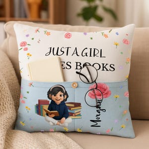 May include: A decorative pillow with a light blue pocket featuring a cartoon girl reading a book, surrounded by stacks of books. The pillow has the text "JUST A GIRL LOVES BOOKS" and a name. A book and glasses are in the pocket.