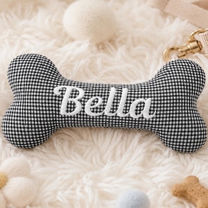 Peut inclure: Un jouet en forme d'os pour chien &agrave; carreaux noirs et blancs avec le nom "Bella" brod&eacute; en &eacute;criture blanche. Le jouet est entour&eacute; d'autres jouets et accessoires pour chiens, dont une balle blanche et une laisse.