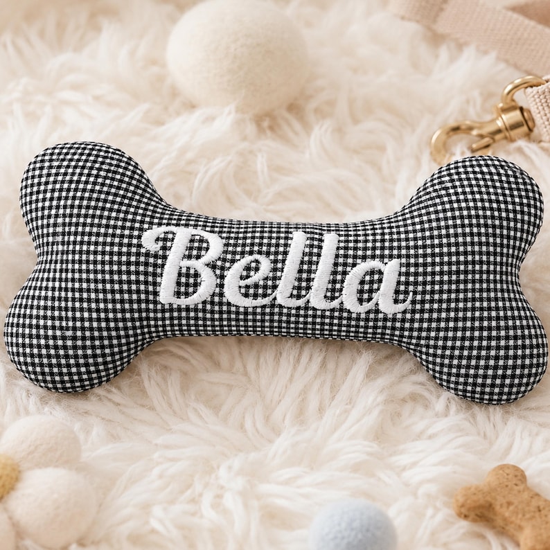 Peut inclure: Un jouet en forme d'os pour chien &agrave; carreaux noirs et blancs avec le nom "Bella" brod&eacute; en &eacute;criture blanche. Le jouet est entour&eacute; d'autres jouets et accessoires pour chiens, dont une balle blanche et une laisse.