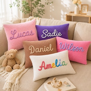 May include: Five rectangular corduroy throw pillows in pink, purple, brown, and white. Each pillow features a unique name embroidered in raised, textured yarn lettering: Lucas, Sadie, Daniel, Wilson, and Amelia. The pillows are arranged together.