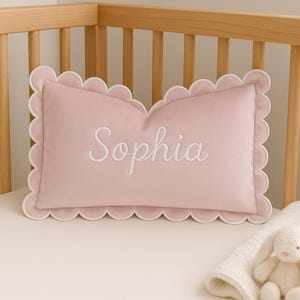 May include: A light pink rectangular pillow with the name "Sophia" embroidered in white script. The pillow has a scalloped edge and is placed on a white surface near a crib.