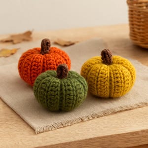 May include: Three small, round, crocheted pumpkins in orange, green, and yellow. Each pumpkin features a thick, ribbed texture and a short, brown, crocheted stem at the top. The pumpkins are arranged in a cluster, showcasing their distinct colors.