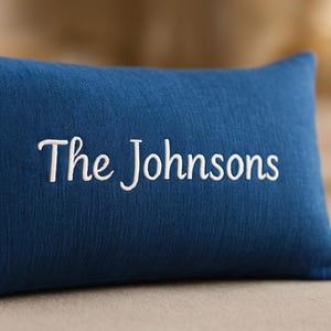 May include: A navy blue rectangular pillow with the embroidered text "The Johnsons" in white script. The pillow has a textured appearance and is likely made of fabric. The background is a blurred neutral tone.
