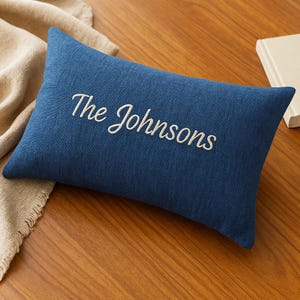 May include: A rectangular, navy blue pillow with the embroidered text "The Johnsons" in a cursive font. The pillow is resting on a wooden surface, with a beige fabric partially visible in the background. A book is also visible.