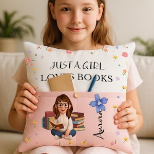 May include: A pink and white pillow with the text "JUST A GIRL LOVES BOOKS" and floral accents. The pillow features a pocket with a cartoon illustration of a girl reading, a notebook, and a pen. The name "Aurora" is written on the pillow.