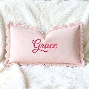 May include: A light pink rectangular pillow with a scalloped edge and the name "Grace" embroidered in a cursive font. The pillow is resting on a white, fluffy surface.