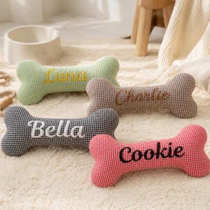 May include: Four dog bone-shaped pillows in green, brown, black, and red gingham patterns. Each pillow has a dog's name embroidered on it: Luna, Charlie, Bella, and Cookie. A white dog bowl is in the background.