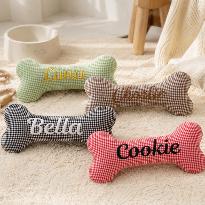 Peut inclure: Quatre coussins pour animaux de compagnie en forme d'os, &agrave; motifs vichy vert, marron, noir et rouge. Chaque coussin porte le nom d'un animal brod&eacute; : Luna, Charlie, Bella et Cookie. Un bol blanc pour chien est en arri&egrave;re-plan.