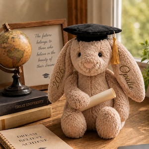 May include: A soft, beige bunny toy wearing a black mortarboard hat with a gold tassel. The bunny holds a scroll and has "2026" and "Opalie" embroidered on its ears. A framed print with text and a globe are in the background.