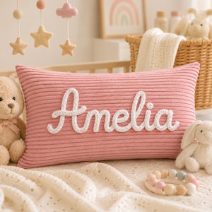 May include: A rectangular pink corduroy pillow featuring the name Amelia in white cursive script lettering. The pillow has a ribbed texture and is positioned in a nursery setting with a teddy bear, a white knitted blanket, and a wooden bead toy.