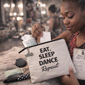 May include: A canvas makeup bag with the text "EAT. SLEEP. DANCE. REPEAT" is held by a person. The bag is open, revealing tissues, a compact mirror, a towel, and a pen. A charm with "2026" hangs from the zipper.