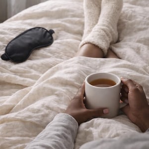 Op de afbeelding: Een persoon in bed houdt een witte mok thee vast. De persoon draagt een grijs shirt met lange mouwen en zachte witte sokken. Een zwart oogmasker ligt op het bed. Het beddengoed is lichtbeige.