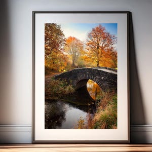 May include: Framed print featuring a stone bridge over a river, set amidst autumnal trees. The artwork showcases the rich colours of autumn, with the bridge's arch mirrored in the water. The scene is set in a natural landscape, evoking a sense of tranquility.