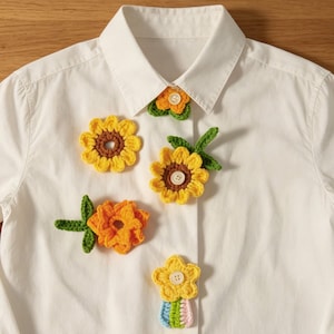 Könnte beinhalten: Ein weißes Hemd mit Knopfleiste, verziert mit mehreren handgefertigten Häkelblumen-Applikationen. Die floralen Designs bestehen aus gelbem, orangefarbenem und grünem Garn, wobei einige Mitten mit kleinen, runden, weißen Kunststoffknöpfen akzentuiert sind.