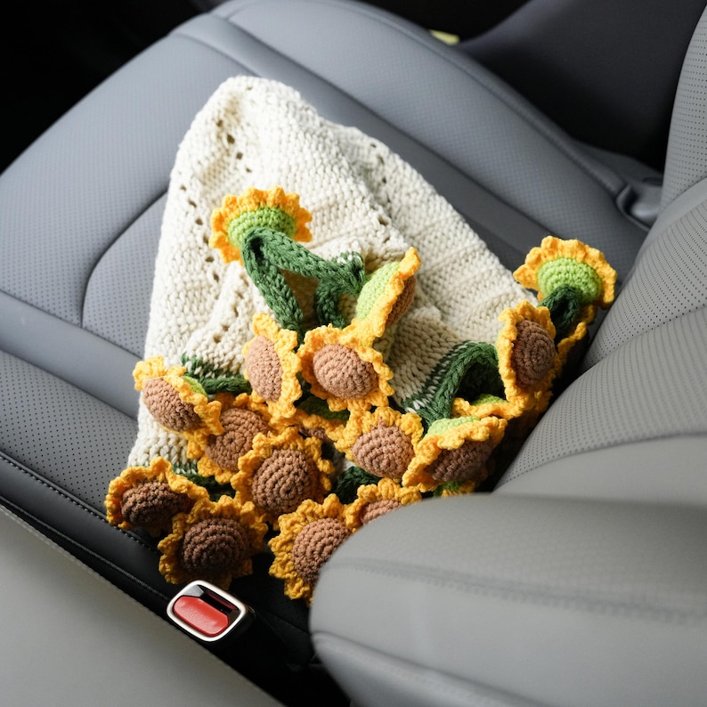 May include: A handmade crochet sunflower garland with yellow petals, brown centers, and green stems, draped over a cream-colored crocheted blanket. The garland is resting on a gray car seat.