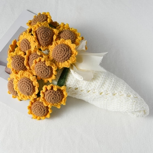 May include: A bouquet of crocheted sunflowers with yellow petals and brown centers. The stems are wrapped in green yarn and tied with a cream-colored ribbon. The bouquet is held in a white knitted sleeve.